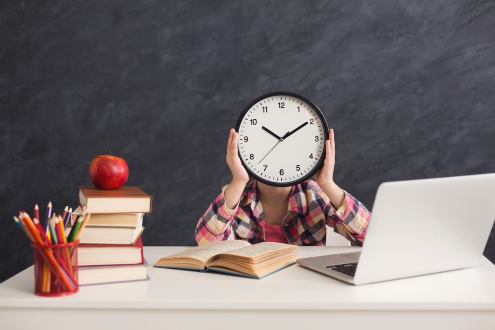 Child sitting at desk holding a clock over their face with open book and computer on the desk.