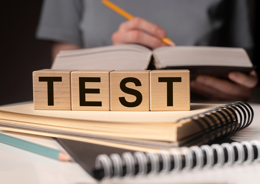 child with book opened and writing with a pencil while wooden blocks spelling "TEST" sit in front.