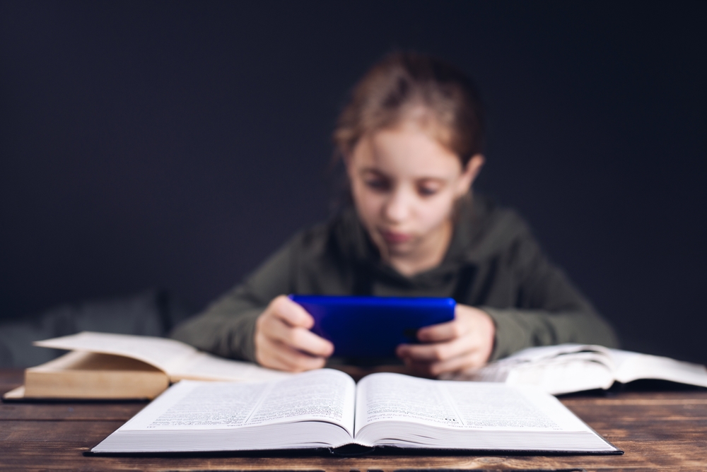 A,Girl,With,A,Phone,In,Her,Hands. Open book on table.